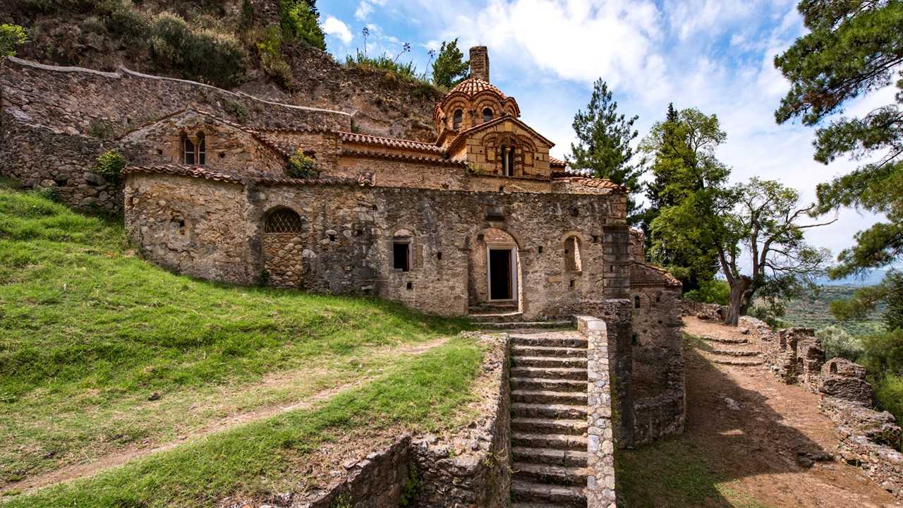 Exterior view of the Perivleptos Monastery Katholikon in Mystras, showing the late Byzantine stone and brick church built into the side of a cliff with its rugged masonry walls, traditional pitched roof, and adjacent tower and monastic buildings under a clear sky, reflecting its 14th-century Mystras architectural style and historic setting on the hillside.
