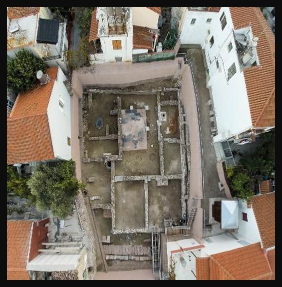 Aerial view of the House of Menander archaeological site in Mytilene, Lesbos, showing the rectangular remains of a late Roman-period peristyle house with visible outlines of walls and courtyard foundations set into the urban fabric.