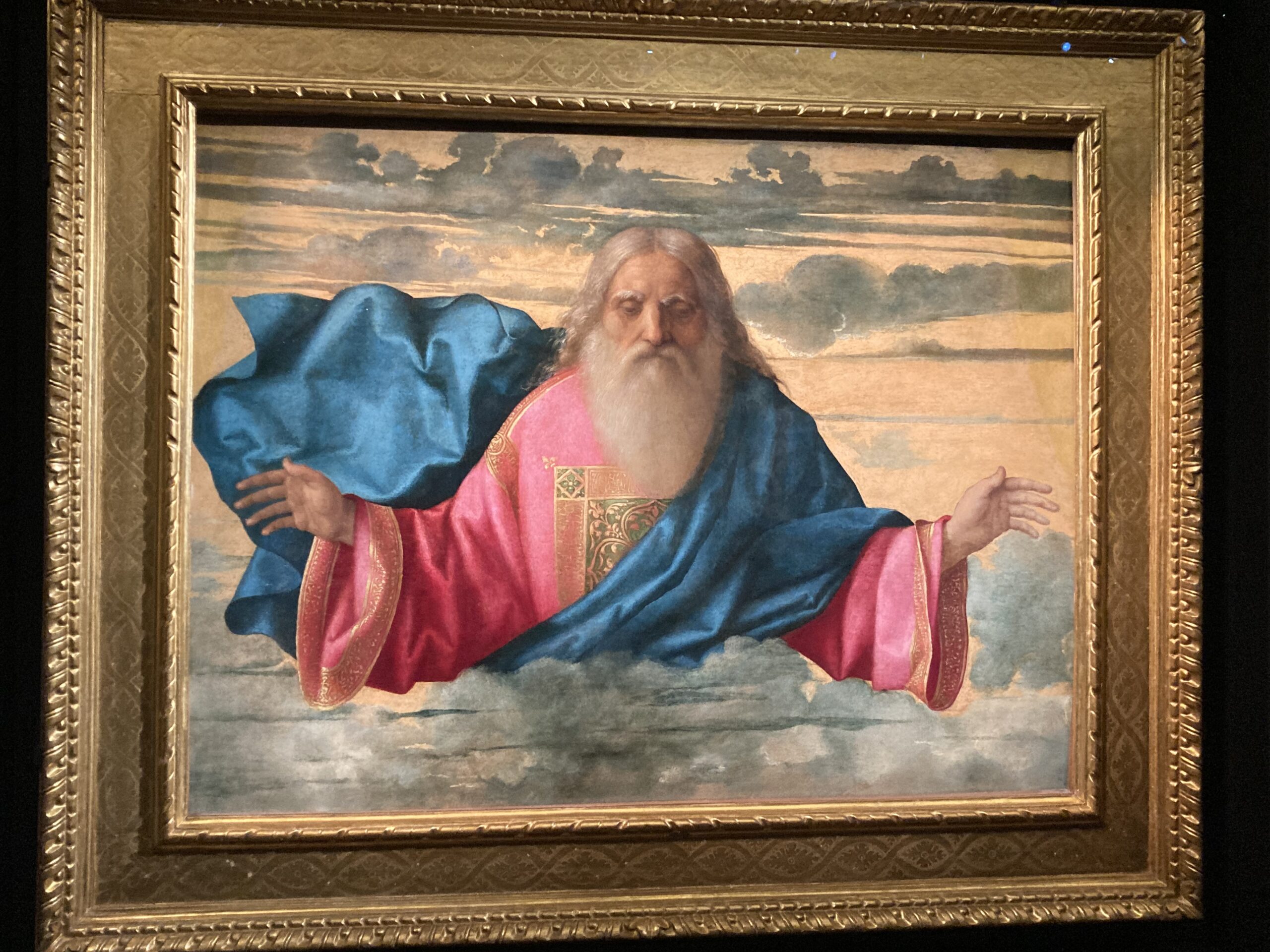 God the Father appears as an elderly, white-bearded man in red and blue robes, shown against glowing clouds and raising his hand in blessing.