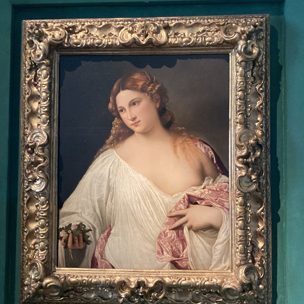 Titian’s Flora, a young woman with long copper-blonde hair wearing a white draped garment, holds a small bouquet of violets, rosebuds and jasmine against a dark background.