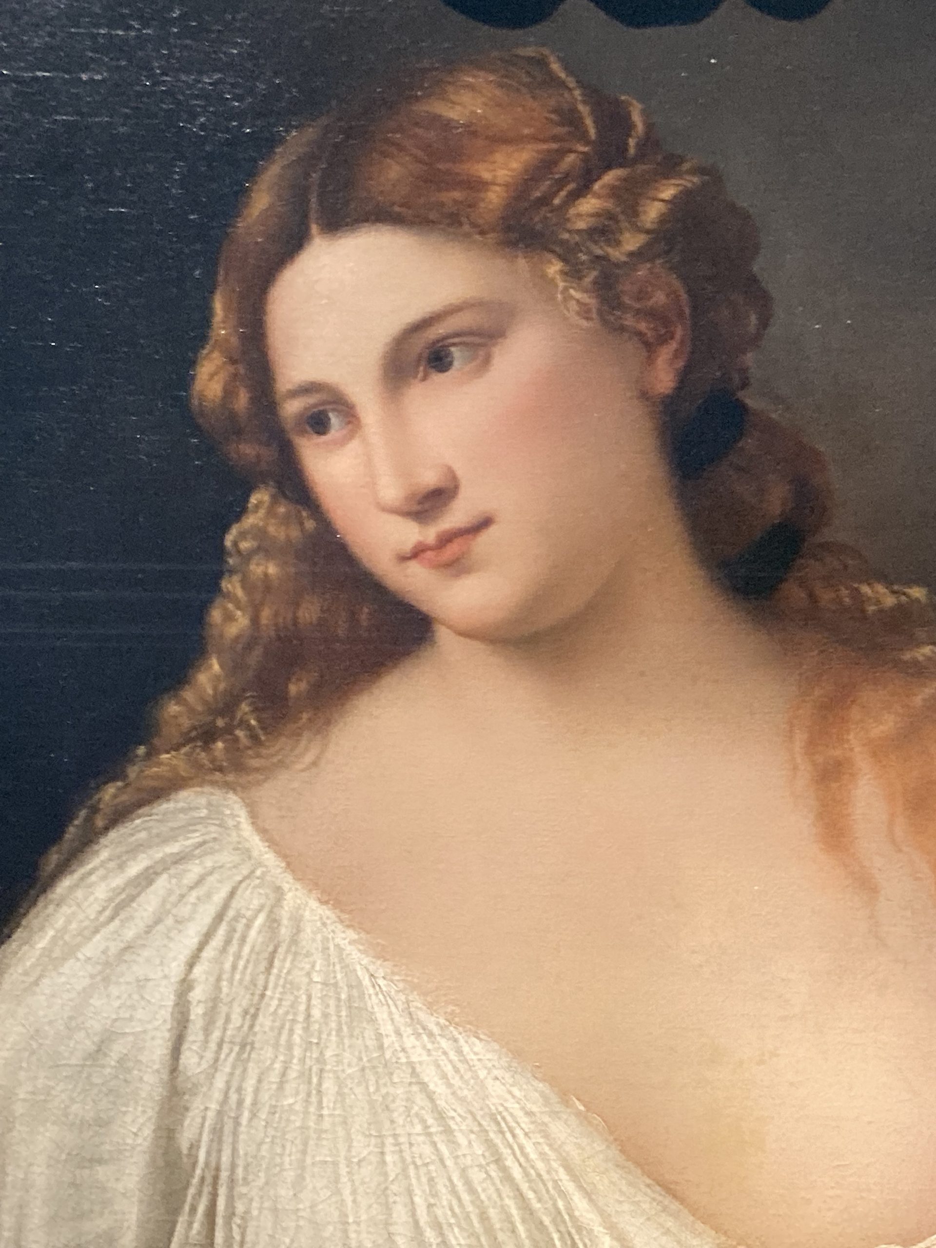 Titian’s Flora (detail: face), a young woman with long copper-blonde hair wearing a white draped garment, holds a small bouquet of violets, rosebuds and jasmine against a dark background.