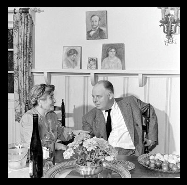Gabrielle Renard and Jean Renoir seated together at a dining table, with flowers, wine, and paintings by Pierrre-Auguste Renoir on the wall behind them.
